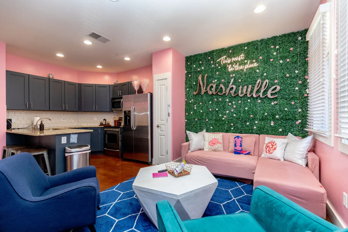 A vibrant living room with a bold green wall adorned by a glowing 'Nashville' sign, a plush pink sectional sofa perfect for groups, chic blue accent chairs, and a trendy geometric coffee table, all set in a playful, music-inspired atmosphere.