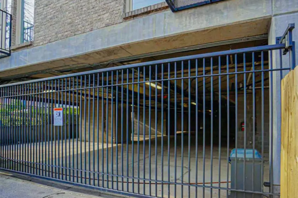 A view of the secure garage entrance, framed by a metal fence and surrounded by concrete walls. The setting conveys safety, privacy, and practicality for parking needs.