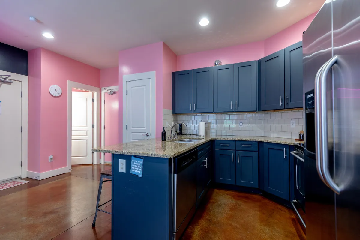 A modern kitchen with pink walls, blue cabinets, a granite countertop, and stainless steel appliances. The space is both playful and functional, inviting guests to cook, entertain, or enjoy a quick coffee before heading out.