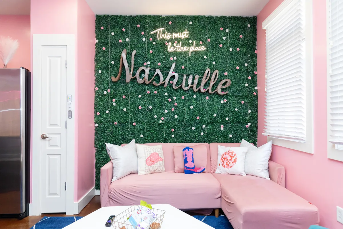 A cheerful, vibrant living room with a pink couch, decorative pillows, and a green wall adorned with flowers. The focal point is a neon sign reading 'Nashville,' making this a perfect backdrop for group selfies. The space is designed for socializing and cozy gatherings, bursting with playful energy and photogenic décor.