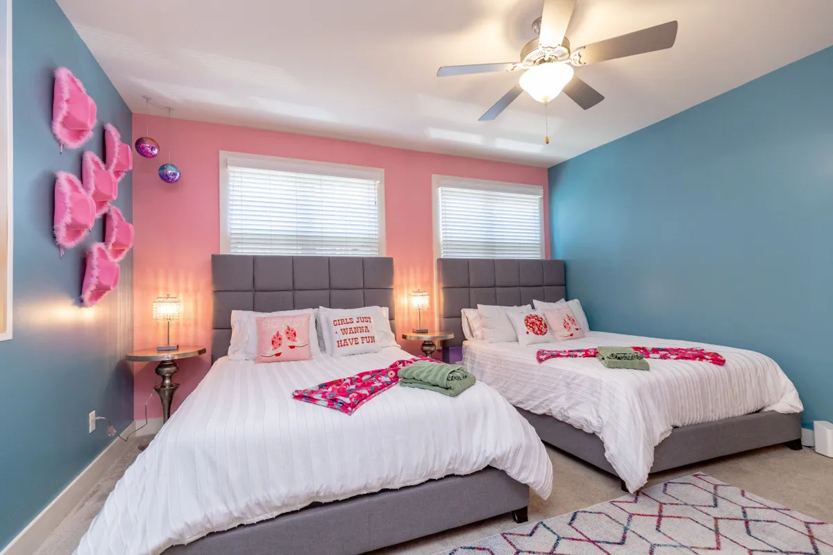 This image captures two large beds adorned with decorative pillows and flanked by bedside lamps, set against colorful wall art. The room looks spacious, bright, and playful, inviting relaxation and group gatherings.