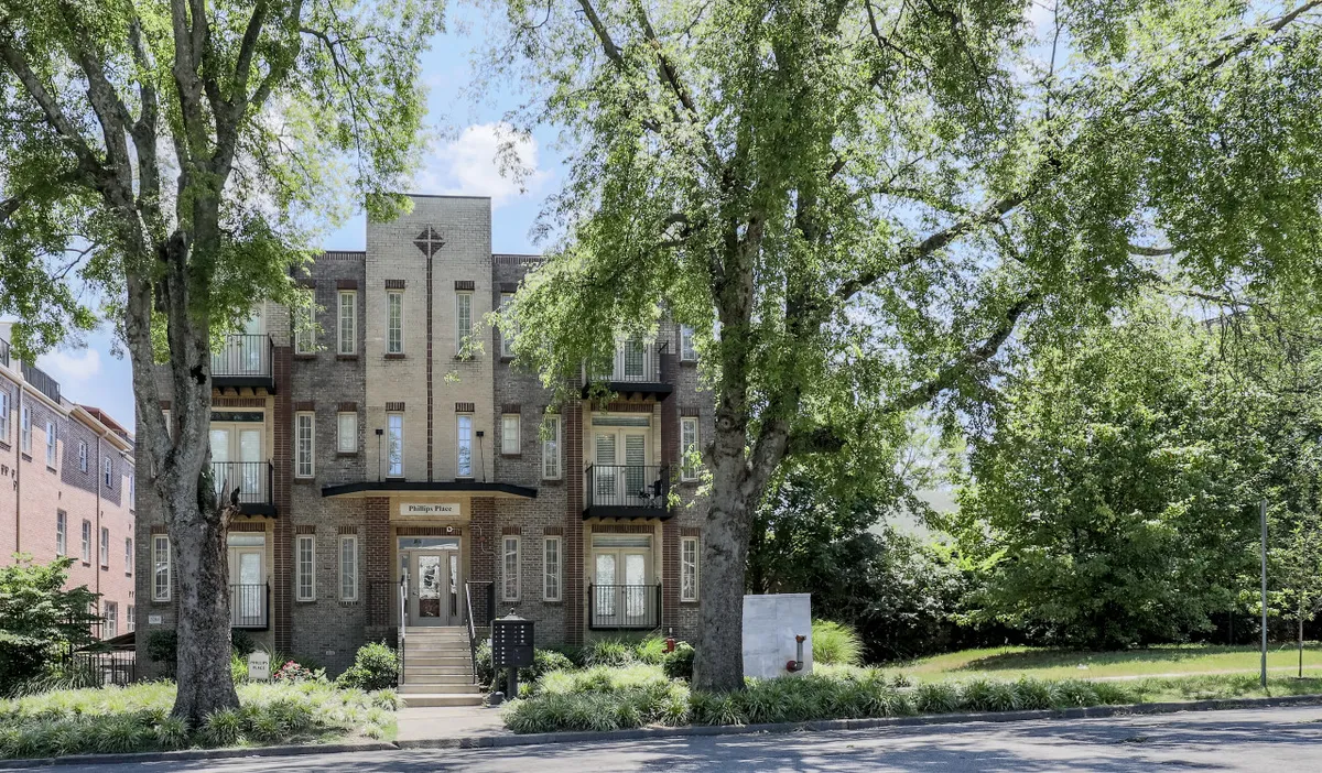 The image depicts the exterior of the building with large trees providing shade, a front entrance, and balconies where guests can gather. The scene feels inviting, suggesting easy access to the broader neighborhood and a relaxing spot for morning coffee or evening chats.