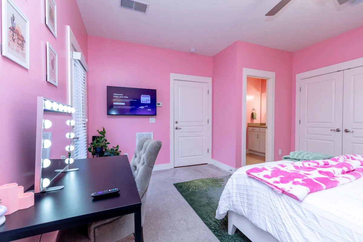 A stylish bedroom featuring a bed with a pink blanket, a vanity mirror with lights, and a television mounted on a pink wall. The setup is both cozy and functional, ideal for getting ready or unwinding after a day out.