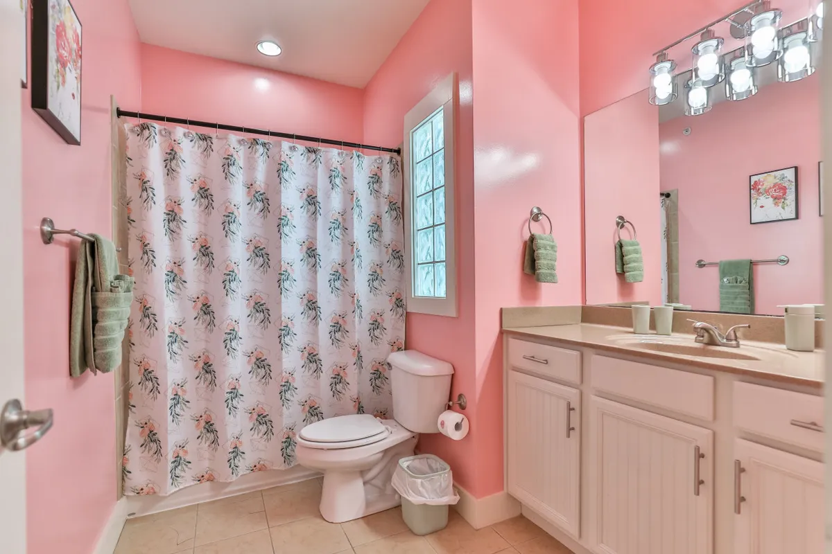 A cheerful bathroom with pink walls, a floral shower curtain, a well-lit vanity, and a window that lets in natural sunlight. The playful decor and functional amenities create a vibrant and refreshing space.